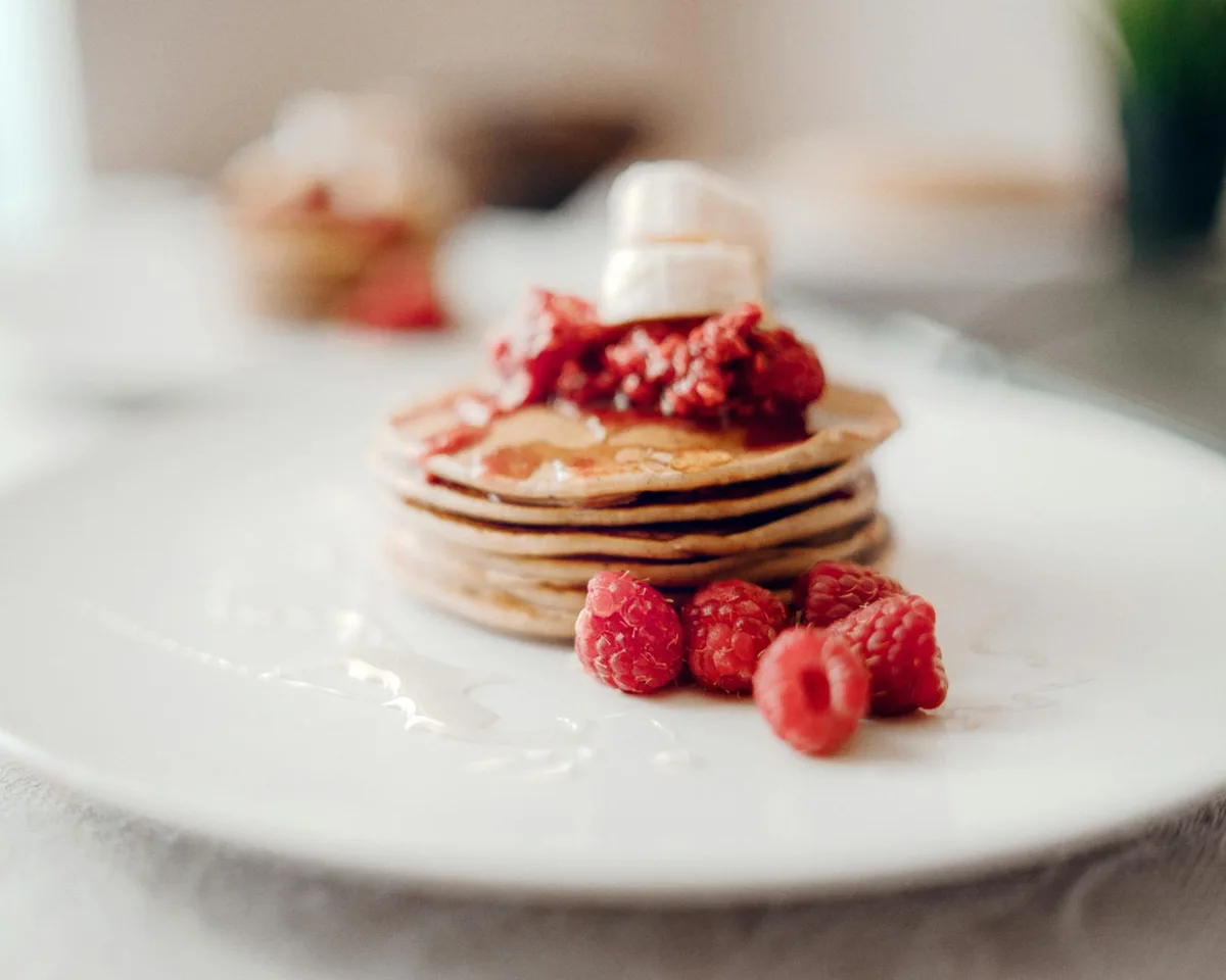 Stack of pancakes topped with fresh raspberries and syrup on a white plate.